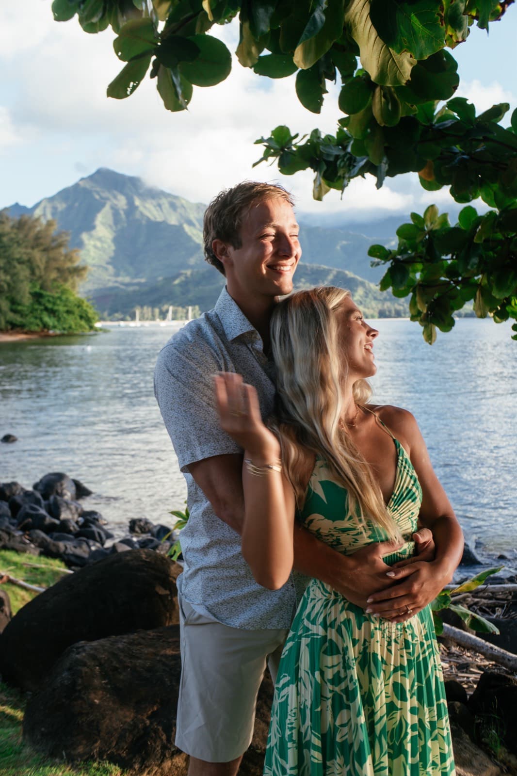 Will hugs Emma from behind, both laughing, by the water with mountains beyond